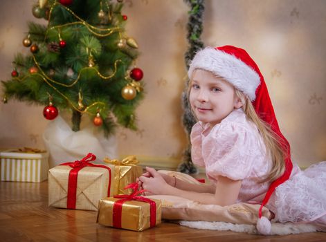 Cute girl in Santa hat dreaming under Christmas tree with gifts