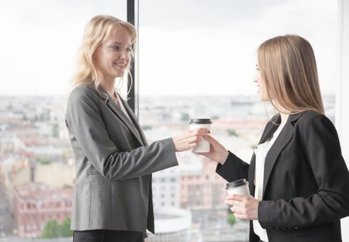 Business woman offering coffee to colleague in office with panoramic windows with view at city