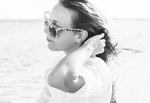 A young woman in sunglasses holds a hand to her hair fluttering in the wind near the sea, monochrome. The concept of a free way of life.