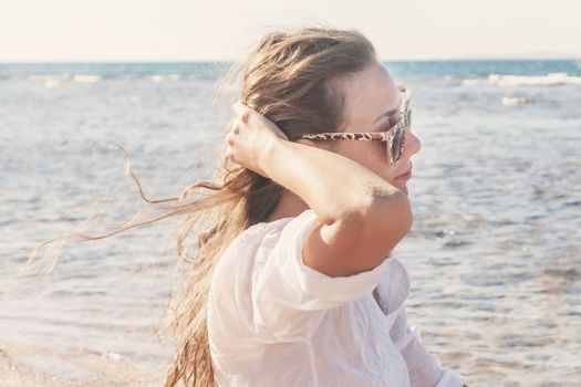 A young woman in sunglasses holds a hand to her hair fluttering in the wind near the sea, toning in retro style. The concept of a free way of life.