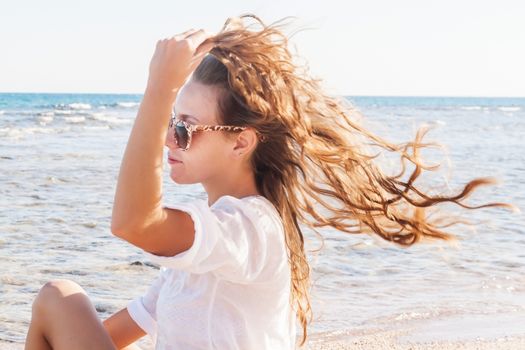 A young woman in sunglasses holds a hand to her hair fluttering in the wind near the sea. The concept of a free way of life.