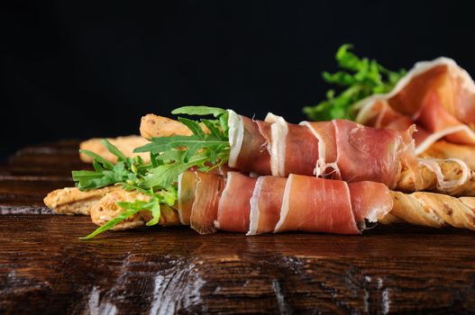 Grissini -   bread sticks with Parmesan, wrapped with a piece of prosciutto and arugula. Italian dish with antipast on a wooden table.