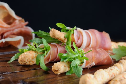 Grissini -   bread sticks with Parmesan, wrapped with a piece of prosciutto and arugula. Italian dish with antipast on a wooden table.