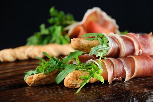 Grissini -   bread sticks with Parmesan, wrapped with a piece of prosciutto and arugula. Italian dish with antipast on a wooden table. 