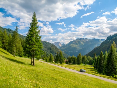 Landscape of Dolomites with green meadows, coniferous trees, blue sky, white clouds and rocky mountains.