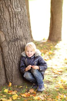 Cute little blond boy sitting near big tree in autumn park and smiling