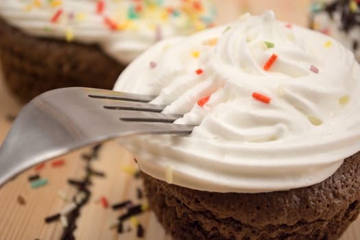 Muffin with cream and colorfull crumbs on top of it sitting on bright wooden table. Fork is pinned in muffin