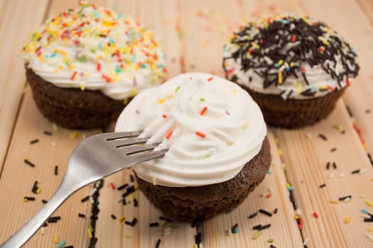 Muffin with cream and chocolate crumbs on top of it sitting on bright wooden table. Fork is pinned in muffin