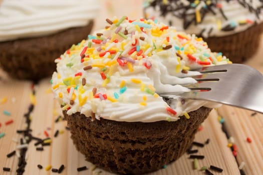 Muffin with cream and colorfull crumbs on top of it sitting on bright wooden table. Fork is pinned in muffin