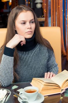 A girl with a book pondered at a table in a cafe, looks out the window