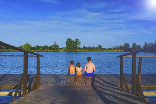 Children sitting on pier. Three children of different age - teenager boy, elementary age boy and preschool girl sitting on a wooden pier. Summer and childhood concept. Children on bench at the lake.