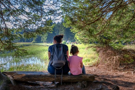 Mother and daughter sitting on bench near mountain lake, facing away, family outdoor hiking in nature, love between mothers and daughters