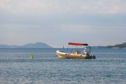 Small rubber motor boat, dinghy in morning light, red sun shade above the boat, moored on buoy, islands in the background
