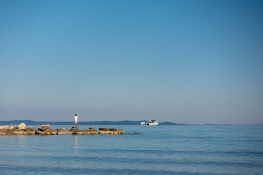 Young boy, facing away, unrecognizable, fishing of stone pier, island in background, copyspace on blue sky background