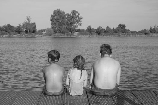 Children sitting on pier. Three children of different age - teenager boy, elementary age boy and preschool girl sitting on a wooden pier. Summer and childhood concept. Children on bench at the lake. Siblings sitting on wooden pier. Black white photo.