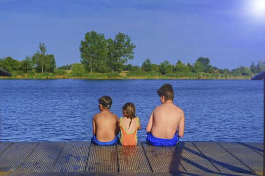 Children sitting on pier. Three children of different age - teenager boy, elementary age boy and preschool girl sitting on a wooden pier. Summer and childhood concept. Children on bench at the lake. Siblings sitting on wooden pier.