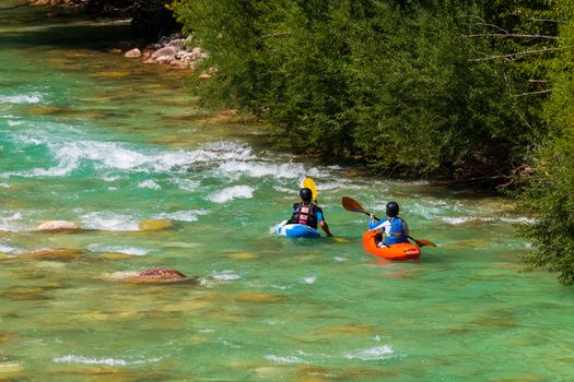 A couple kayaking in emerald, turquoise mountain river, approaching rapids in front of them, wearing wetsuits and avoiding roks and boulders in the streaming water, facing away, red and blue kayak