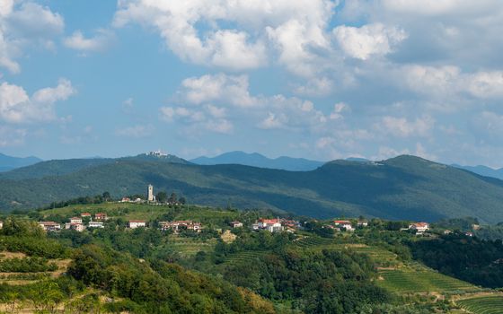 Village of Kojsko, Sloveniain famous wine growing region of Goriska Brda, with vineyards and orchards, lit by sun and clouds in background, holy mountain with church above Nova Gorica in background