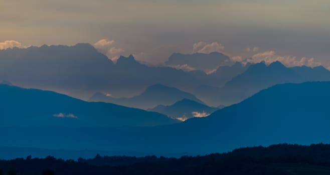 Dolomites in the morning with clouds above, sun rays illuminate the farthest mountain peaks, long distance panoramic shot