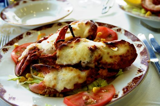 Delicious Grilled Lobster Covered with Fried Cheese on Vegetables Saute Serving on Restaurant Table with Silver Fork and Knife closeup