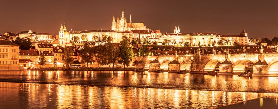 Golden Prague by night. Prague Castle and Charles Bridge reflected in Vltava River. View from Smetana Embankment. Praha, Czech Republic.