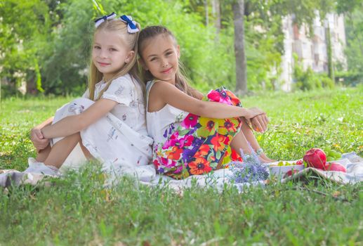 Two smiling girls sitting back-to-back on green grass in the park in summer