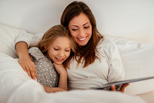 Beautiful smiling mother and her daughter reading book in bed. 