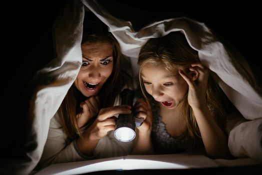 Scaring mother and her daughter reading book under bed cover and holding a flashlight. 