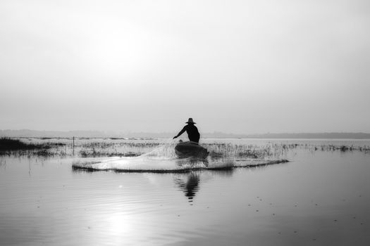 Black and white of fishermen casting for catching the fish on the wooden boat at the lake in the morning. Thailand lifestyle.