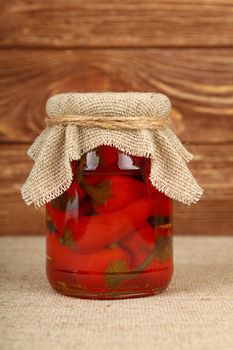 Close up of one glass jar of pickled small red hot cherry chili peppers with canvas top decoration and twine on canvas tablecloth over brown wooden background, low angle side view