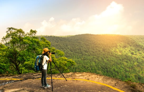 Young traveling woman with backpack hat and camera on tripod stand on the top of the mountain cliff watching beautiful view of woods and sky on her vacation. Asian woman travel alone.