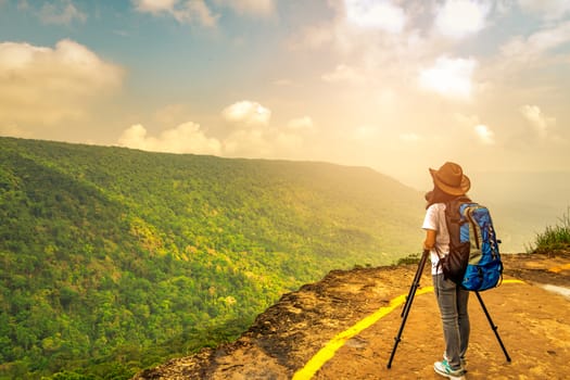 Young traveling woman with backpack hat and camera on tripod stand on the top of the mountain cliff watching beautiful view of woods and sky on her vacation. Asian woman travel alone.