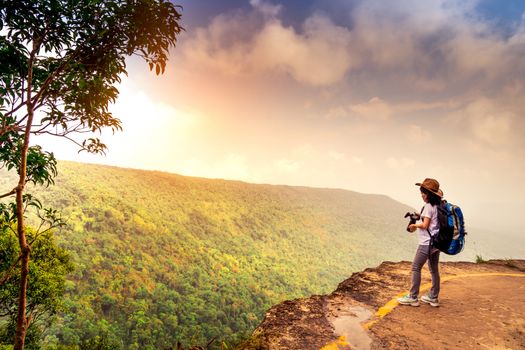 Young traveling woman with backpack hat and camera stand on the top of the mountain cliff watching beautiful view of woods and sky after rain on her vacation. Asian woman travel alone.