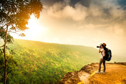 Young traveling woman with backpack, hat and camera stand on the mountain cliff. Female photographer taking picture of tropical forest, sky and clouds on her vacation. Asian woman travel alone.