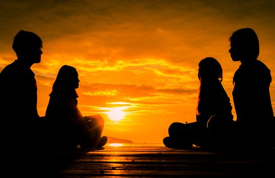 Four young people sit on wooden pier at sunrise on the beach to make meditation with orange  beautiful sky and clouds.