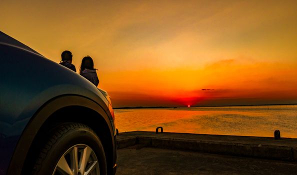 Silhouette of happiness couple standing and relaxing on the beach in front of the car with orange and blue sky at sunset. Summer vacation and travel concept. Romantic young couple dating at seaside.