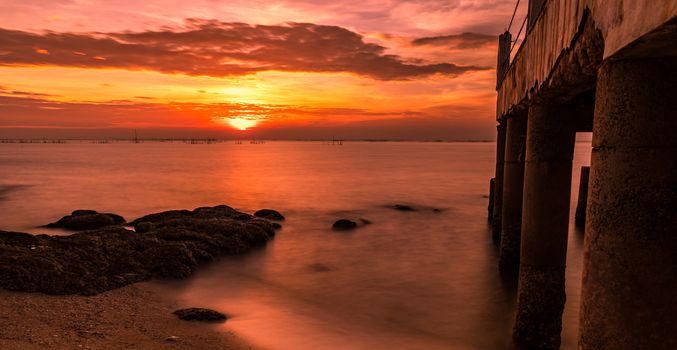 A long exposure landscape of beautiful sunset at the sea beach near concrete bridge with orange sky, clouds and stone