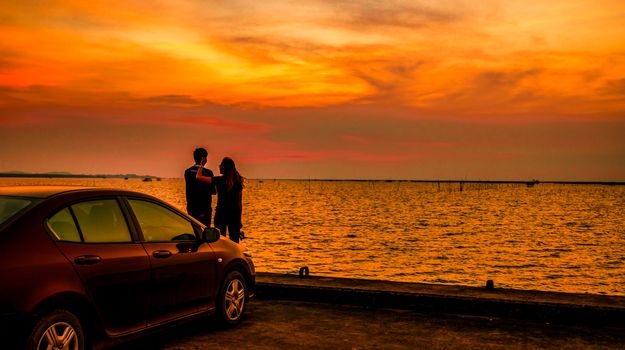 Silhouette of happiness couple standing by the car at the seaside at sunset. Beautiful orange sky and clouds. Road trip.