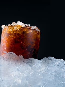 Soft drink with crushed ice cubes in glass put on pile of ice cubes on dark background with copy space. There is a drop of water on the transparent glass surface.