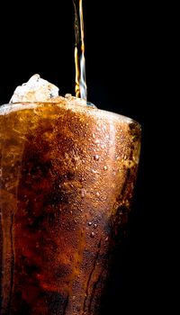 Soft drink pouring to glass with crushed ice cubes isolated on dark background with copy space. There is a drop of water on the transparent glass surface.