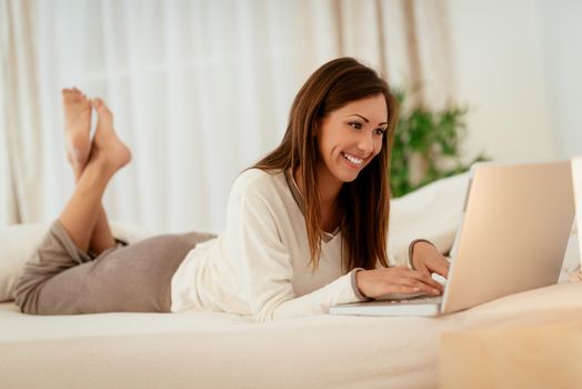 Beautiful smiling girl relaxing in bed and surfing the net on laptop. 