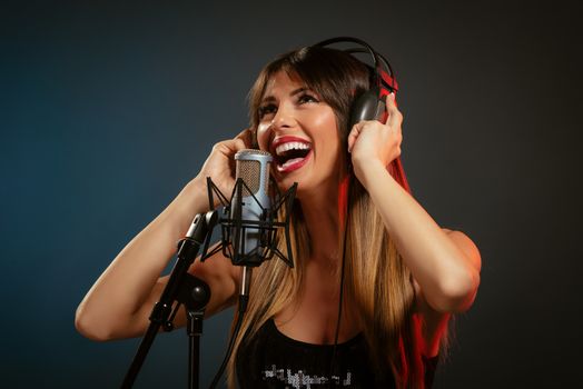 Portrait of a young woman singer with headphones in front of the microphone. Sing with mouth wide open and with an expression of happiness on her face.