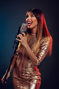 A young smiling woman in sequin dress singing in front of a microphone with expression of happiness on her face.