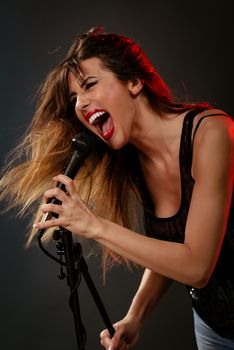 A young woman rock singer with tousled long hair holding a microphone with stand and sing with a wide open mouth.