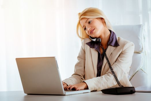 A young beautiful businesswoman sitting in the office, phoning and using a laptop.