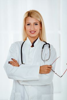 Beautiful young cute female doctor in a white coat standing in the office with a stethoscope over the neck. Looking at camera.