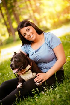 Beautiful young woman and her cute stafford terrier sitting on grass in the park and enjoying. Looking at cemera.