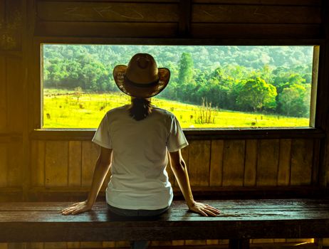 Young Asian woman wear the hat sit on wooden bench and watching beautiful view of green grass field and forest at wildlife observation tower in the evening with warming sunlight. Travel concept.