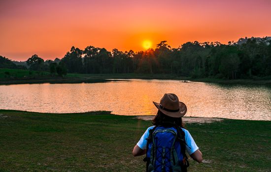 Woman tourist with hat and backpack standing with relax mood and watching sunset behind the pond and evergreen forest. Alone young woman traveller enjoy beautiful scenery sunset.