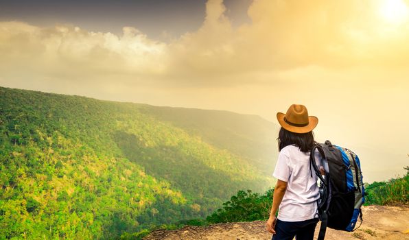 Young traveling woman with backpack and hat stand on the top of the mountain cliff watching beautiful view of woods and sky after rain on her vacation. Asian woman travel alone.